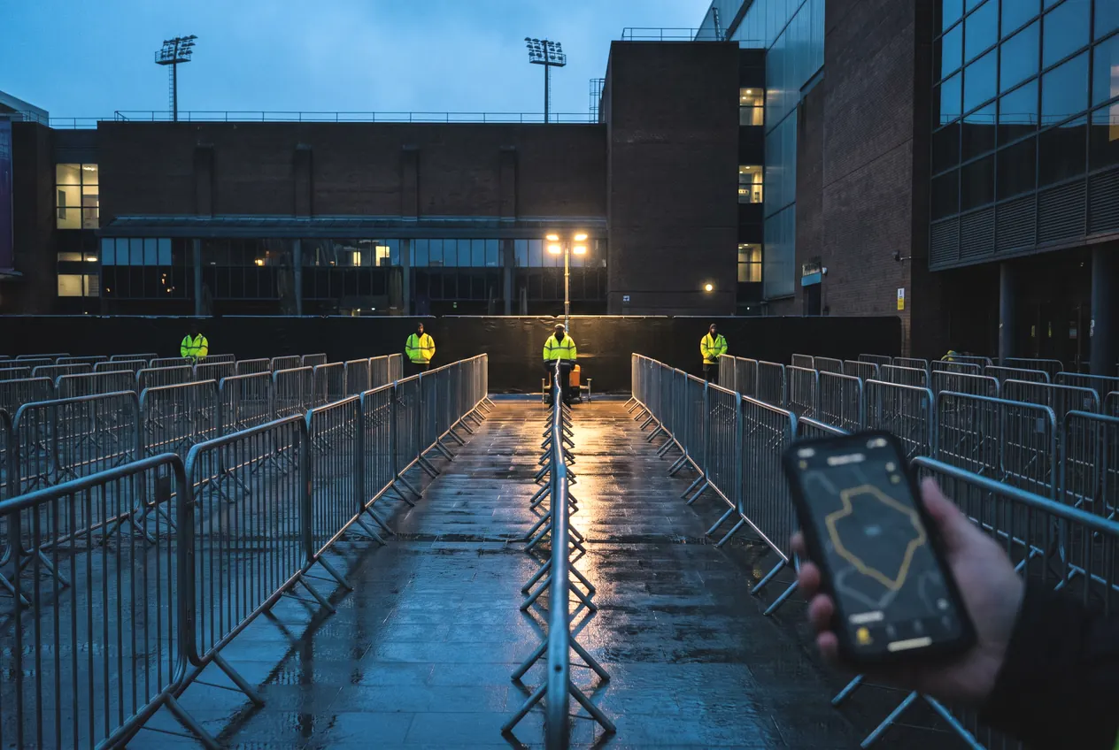 Barrier lanes running up to a floodlit stadium at dusk, a zone map open on a phone in the foreground