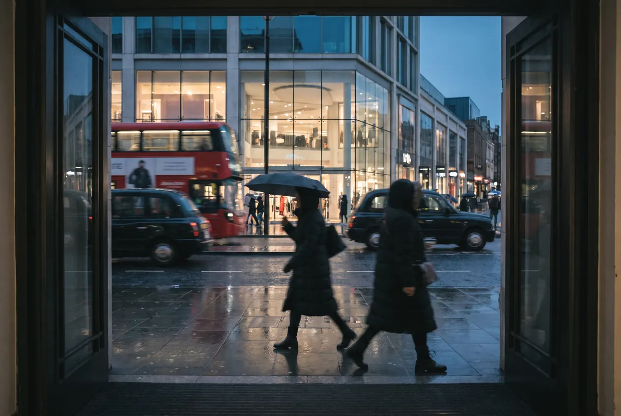 Shoppers passing a store entrance on a wet London street at dusk