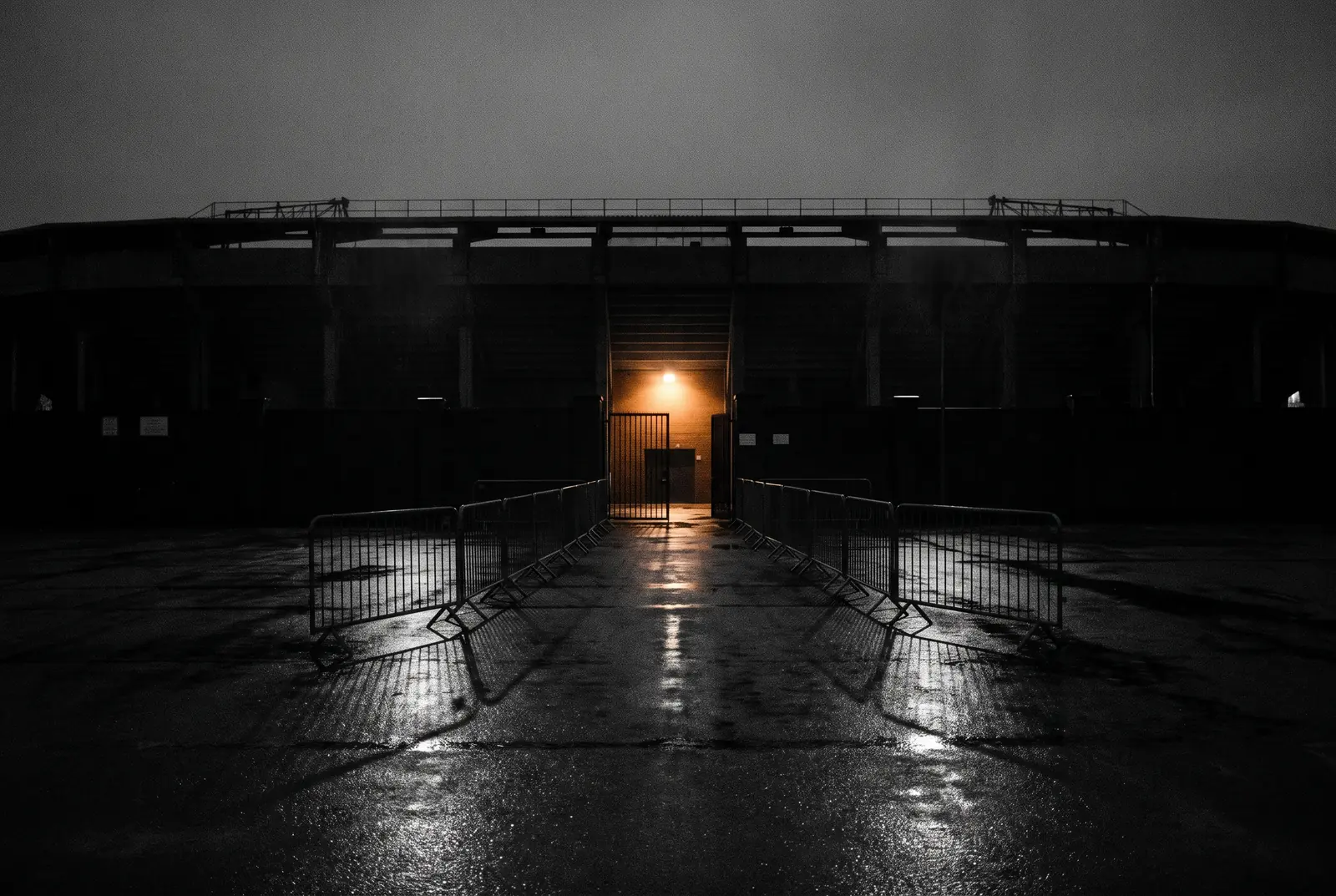 A stadium gate at night behind crowd barriers, one lit doorway in the dark stand