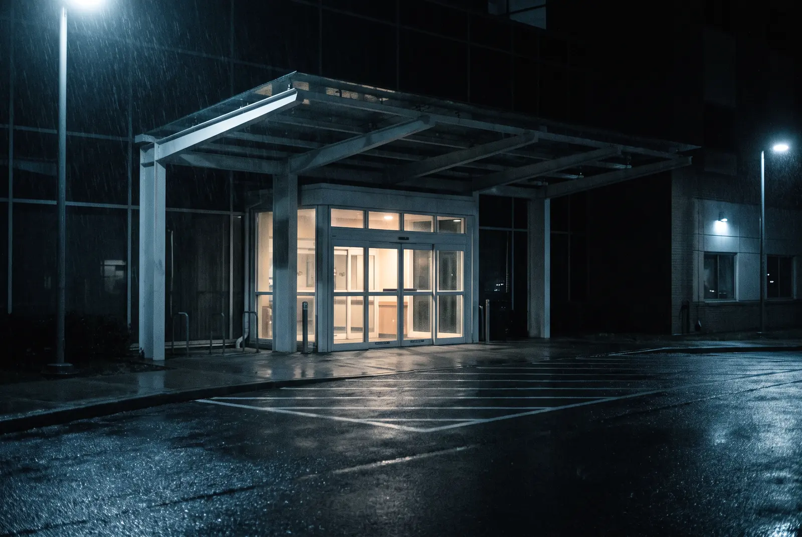 A lit hospital entrance under its canopy in the rain, the forecourt empty at night
