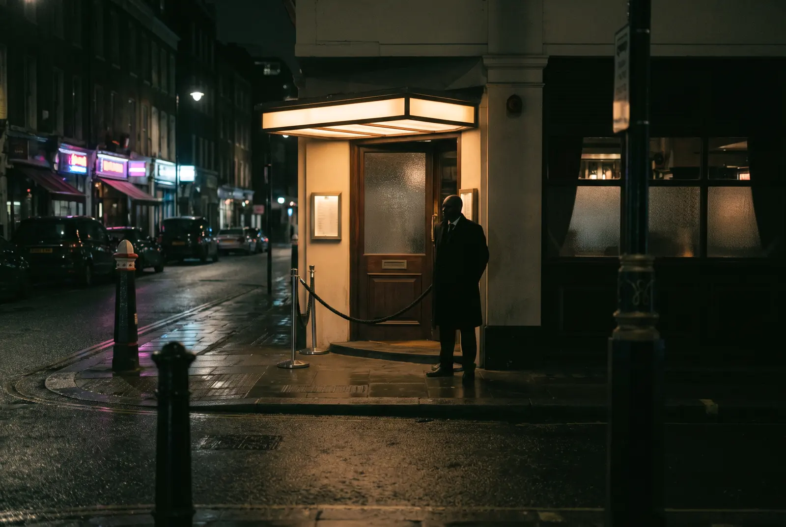 A door supervisor standing under the lit entrance canopy of a bar on a wet city street at night